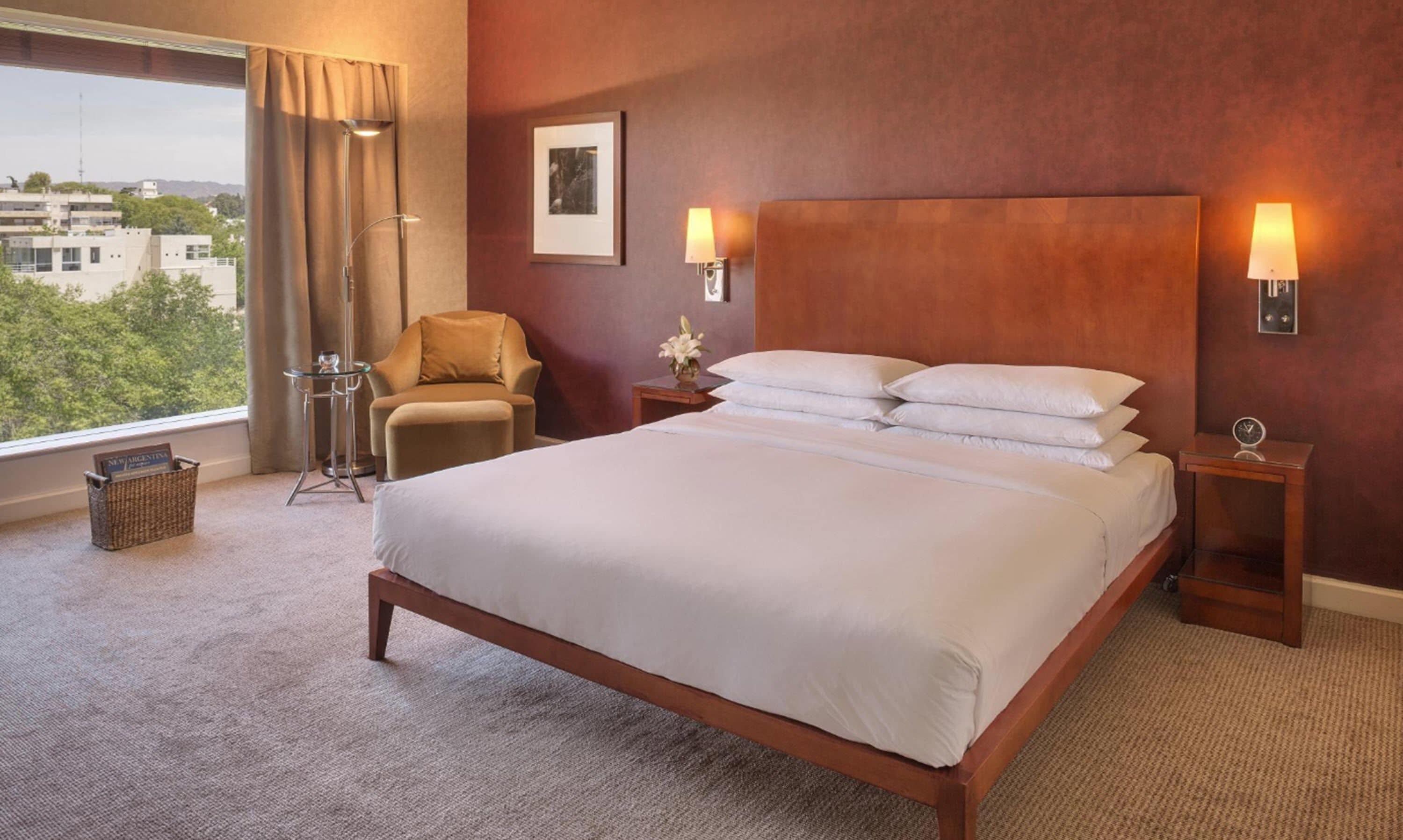 A hotel room with a large bed, a chair, a small table, and a nightstand with a lamp.
