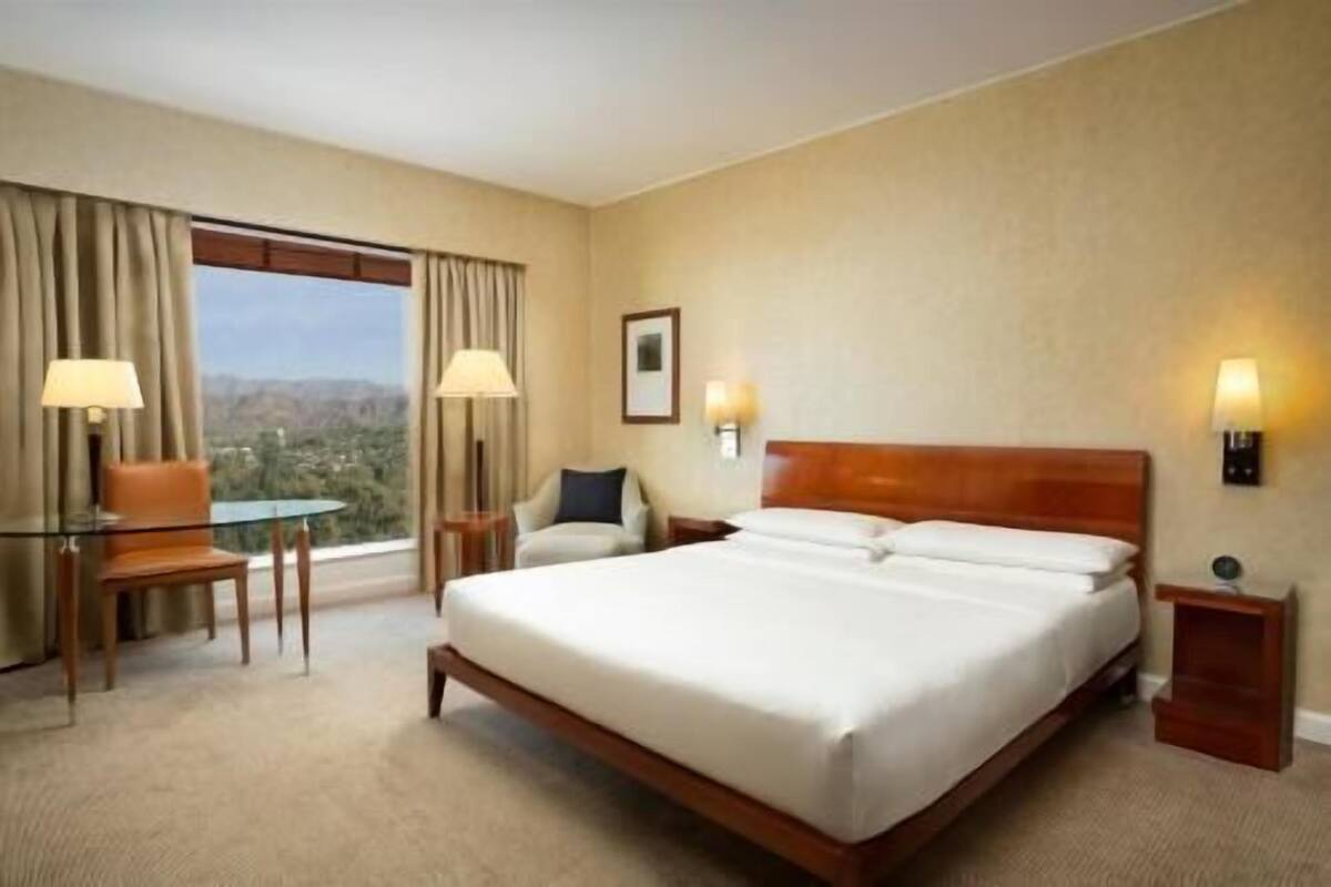 Une chambre d’hôtel avec un grand lit, deux lampes de chevet, une chaise, une petite table et une fenêtre avec des rideaux.