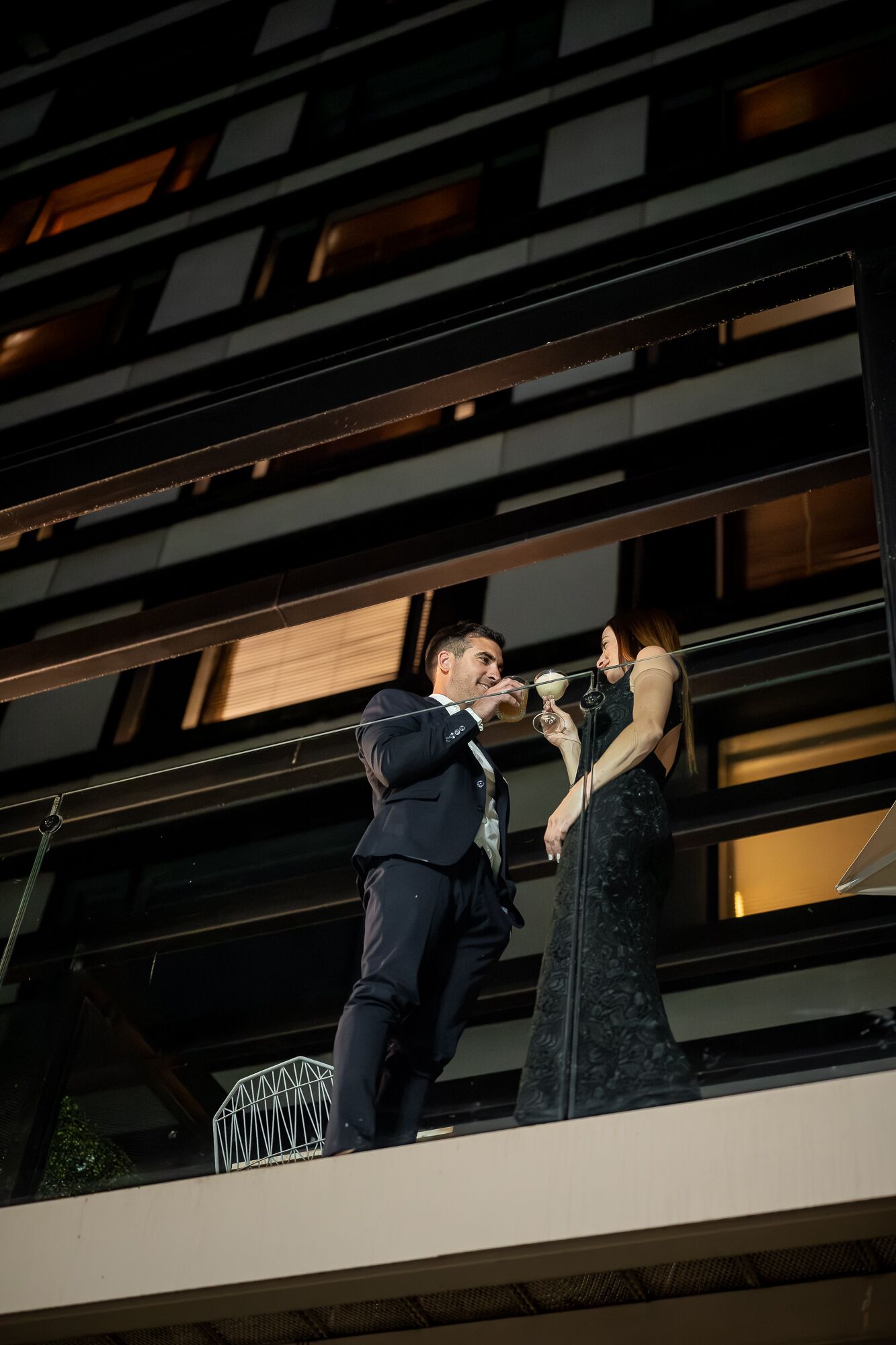 Un hombre y una mujer en un balcón con un edificio moderno al fondo.