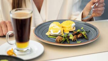 A dining table with a plate of eggs benedict, a glass of beer, and a plate of food in the background.