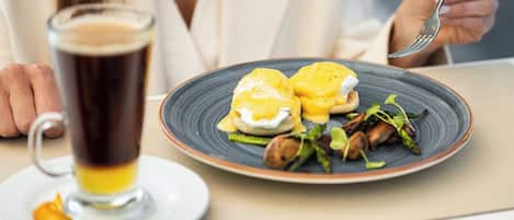 A dining table with a plate of eggs benedict, a glass of beer, and a plate of food in the background.