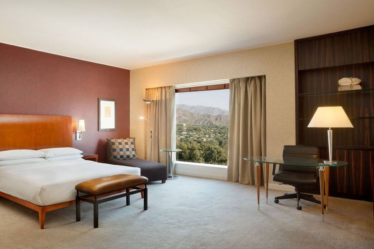Une chambre d’hôtel avec un grand lit, un bureau, une chaise, un banc, une lampe et une vue sur les montagnes.