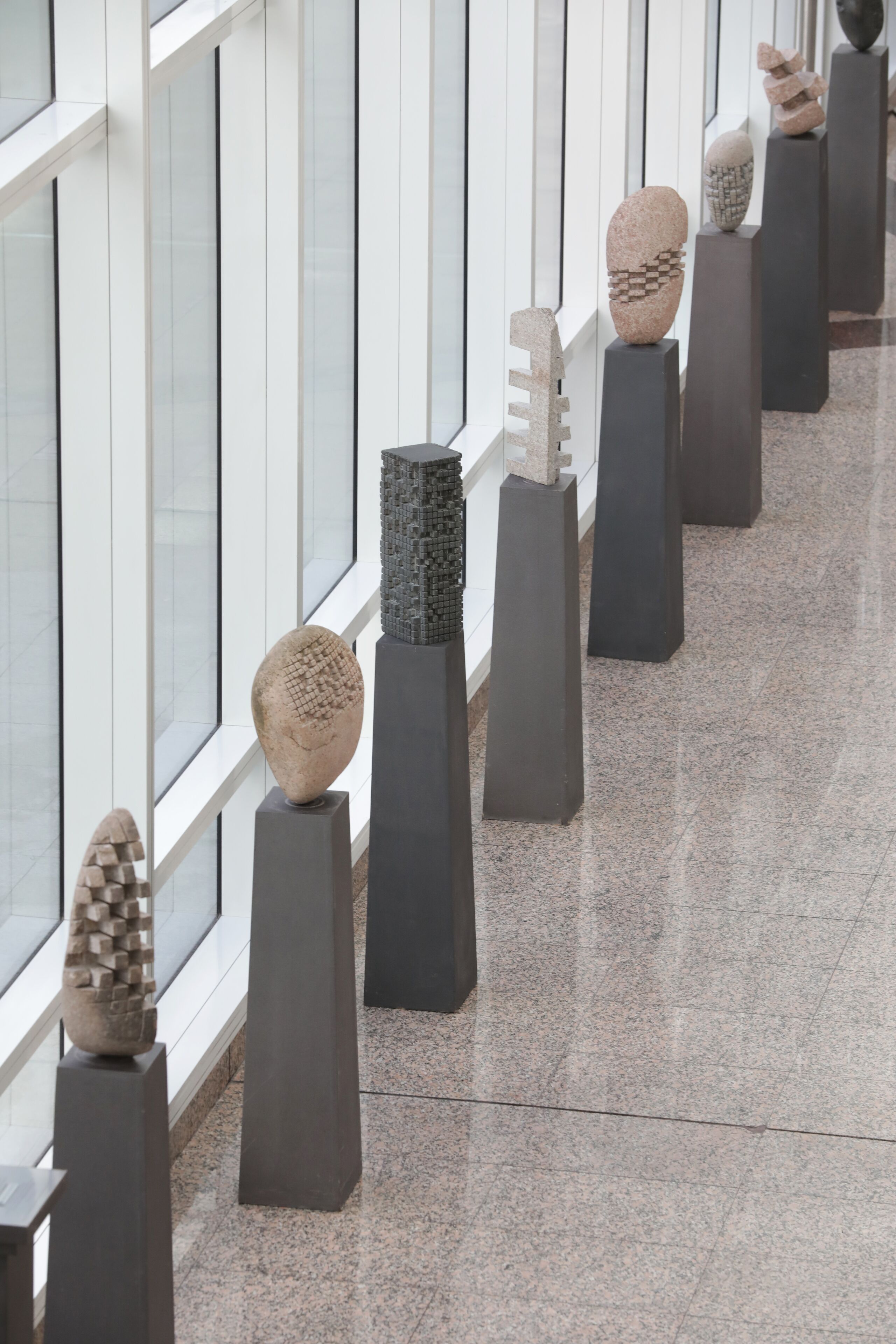 A row of abstract sculptures on pedestals in a modern indoor space with large windows.