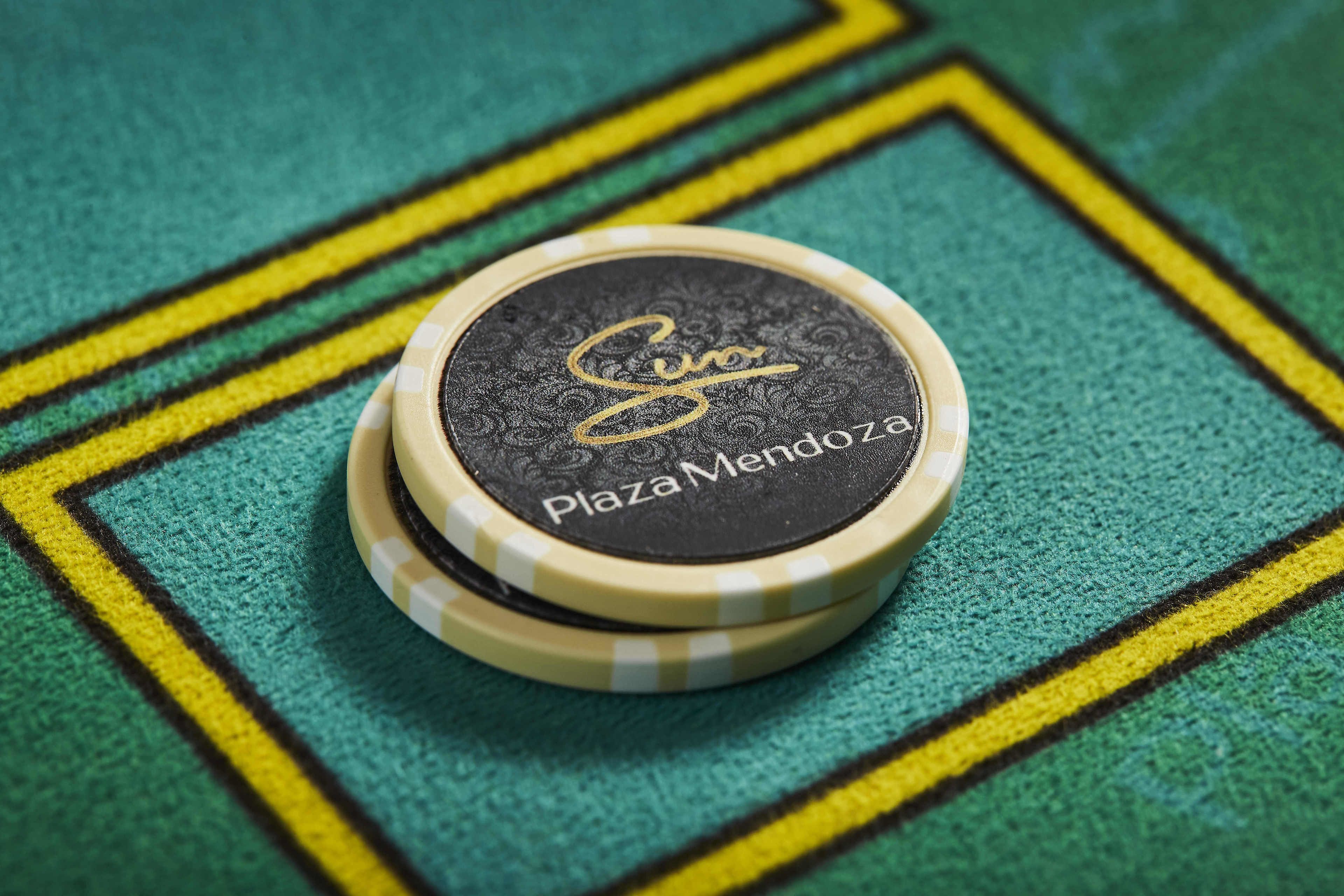 A stack of poker chips with the text "Plaza Mendoza" on them, placed on a green textured surface with yellow lines forming a square pattern.