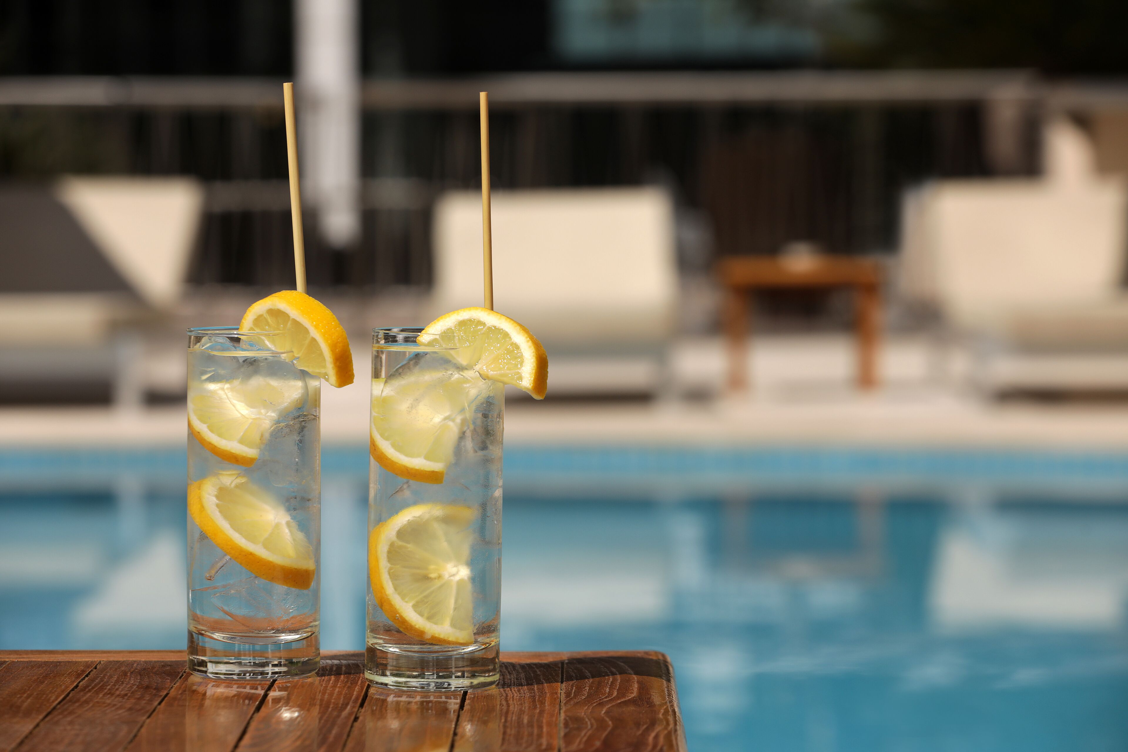 Dos vasos altos de líquido transparente con rodajas de limón y pajitas sobre una mesa de madera junto a una piscina.