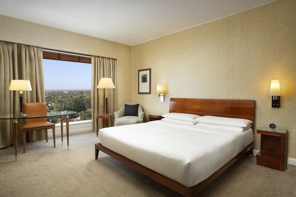 Une chambre d’hôtel avec un grand lit, une table en verre, deux chaises, une vue sur la ville et une table de chevet avec une lampe.