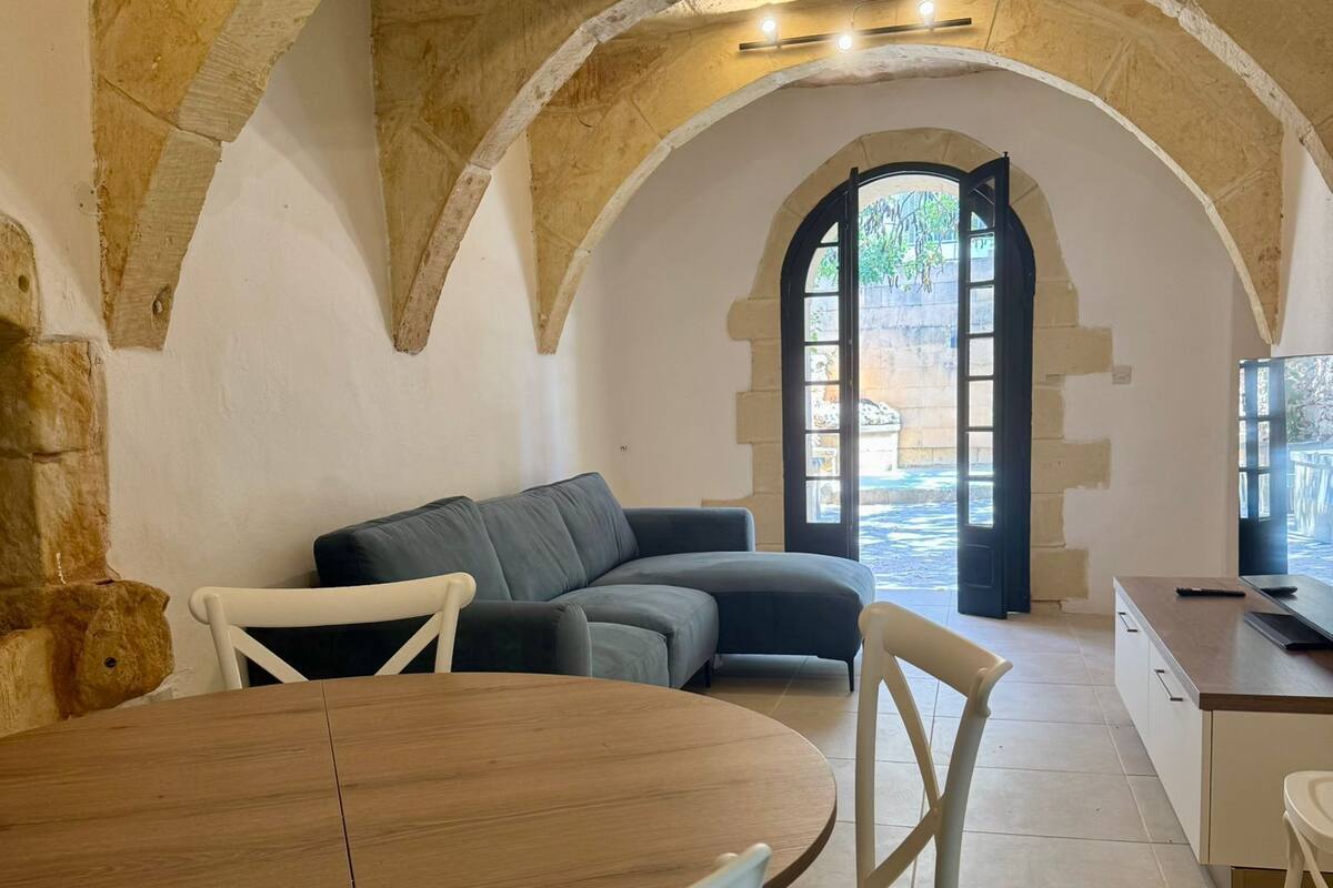 A cozy living area with a stone arch ceiling, a sofa, a dining table with chairs, and a view through an arched doorway to an outdoor space.