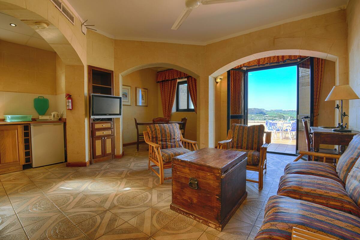 A hotel room with a sofa, chairs, a wooden chest, a television, and a view of the outdoors.