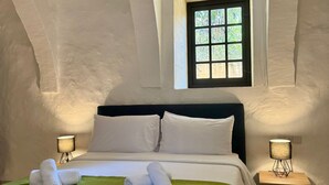 Une chambre à coucher avec un plafond voûté, un grand lit recouvert de linge de lit blanc, deux lampes de chevet et une fenêtre donnant sur un paysage verdoyant.