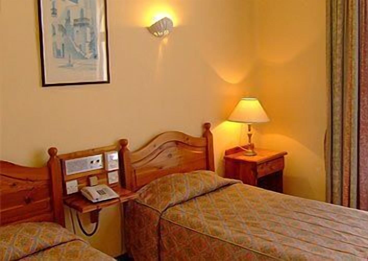 Habitación de hotel con dos camas individuales, cabecera de madera, mesita de noche con lámpara y una lámpara de pared.