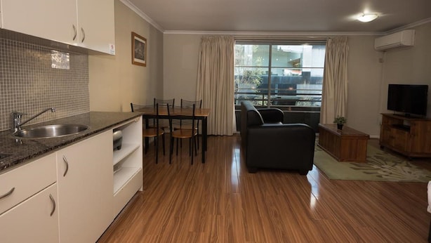 A modern kitchen with a granite countertop and a sink, a dining area with a table and chairs, a dark-colored sofa, and a television on a wooden cabinet.