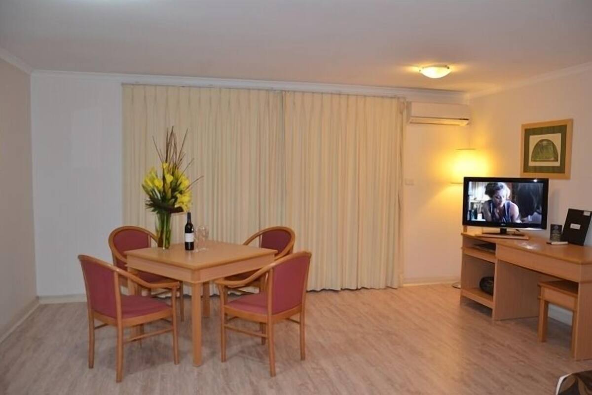 A hotel room with a wooden dining table, two chairs, a television, a small desk, and a curtain.