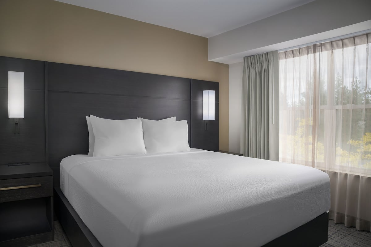 A modern hotel room with a large bed, two bedside tables, and a window with sheer curtains.