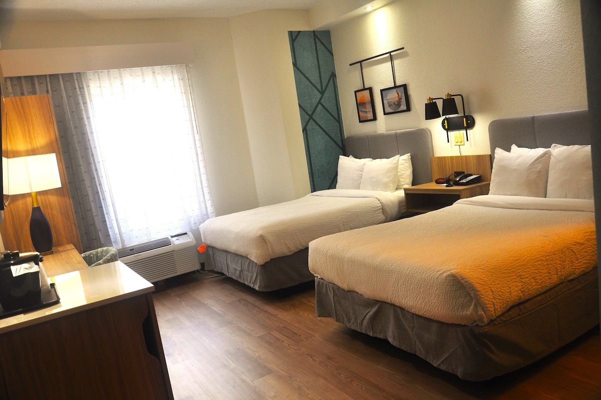 A hotel room with two beds, a desk, a lamp, a heater, and a window with curtains.