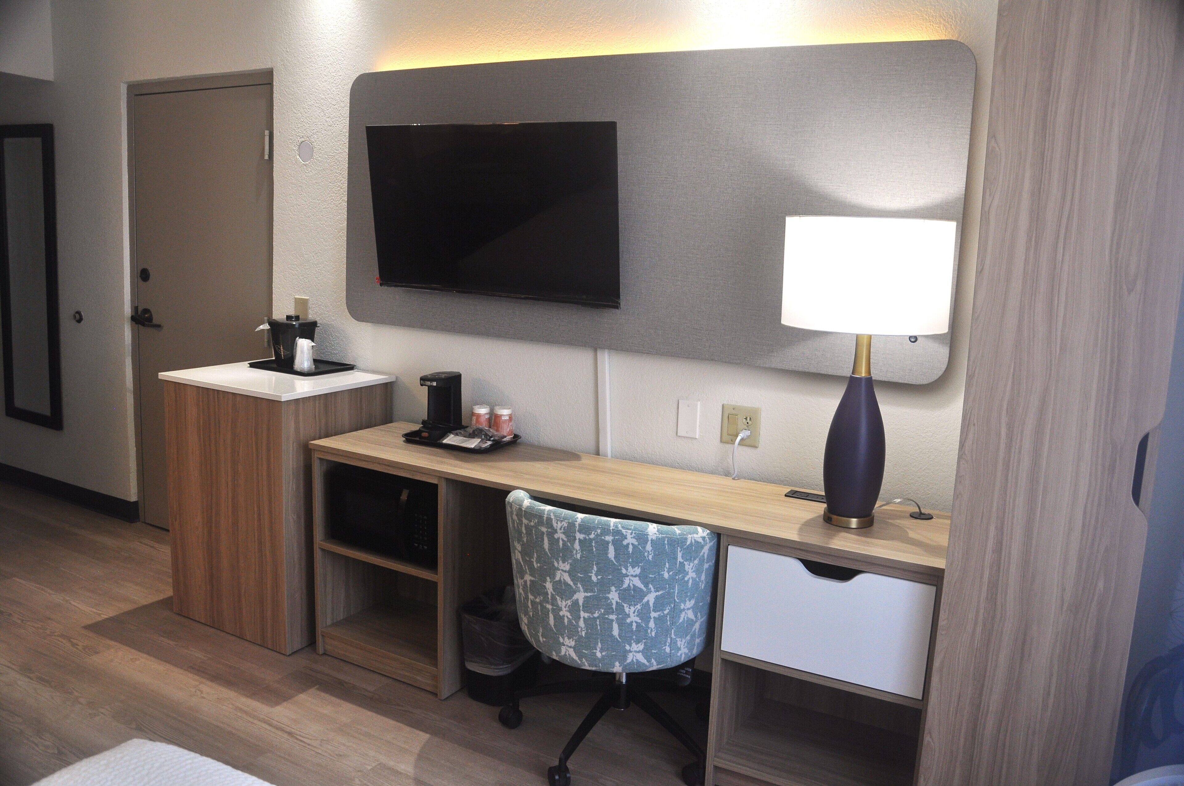 A hotel room with a desk, chair, TV, and a lamp.