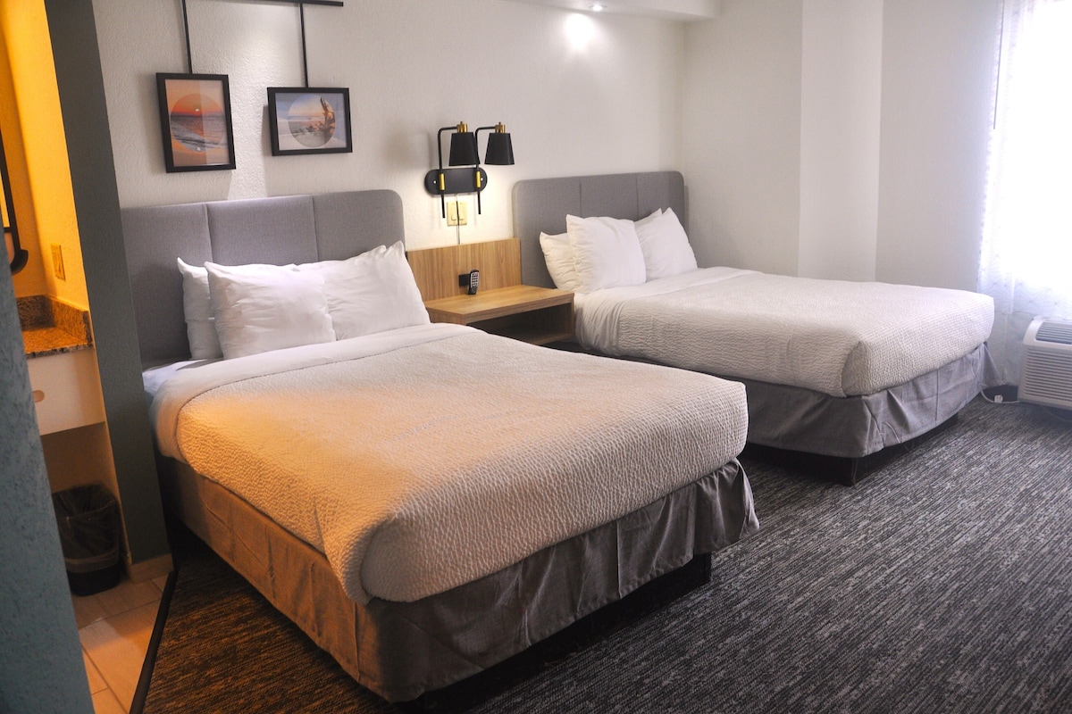 A hotel room with two beds, a nightstand, a lamp, and two framed pictures on the wall.