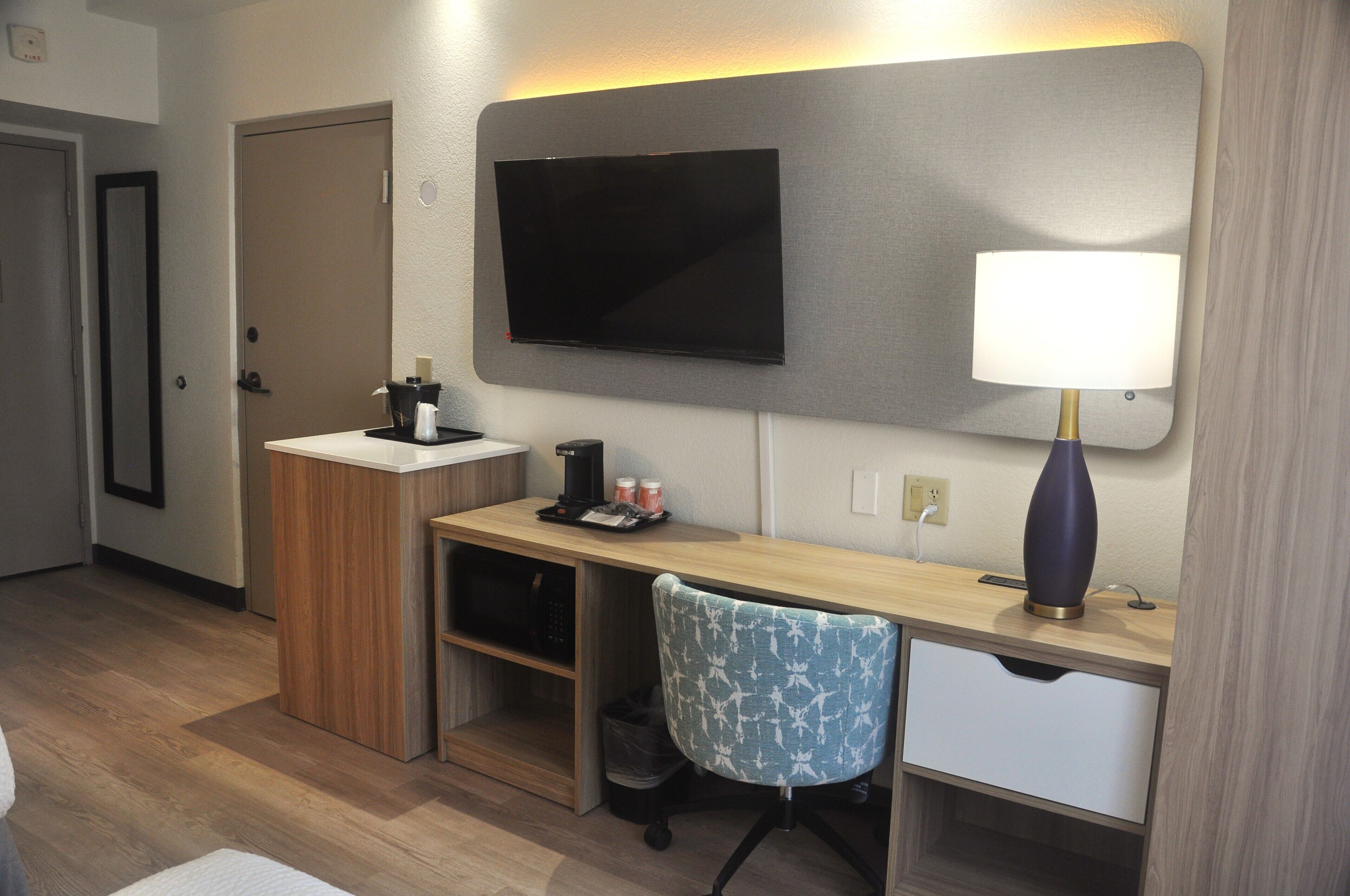 A hotel room with a wooden desk, a chair, a television mounted on the wall, and a bedside lamp.
