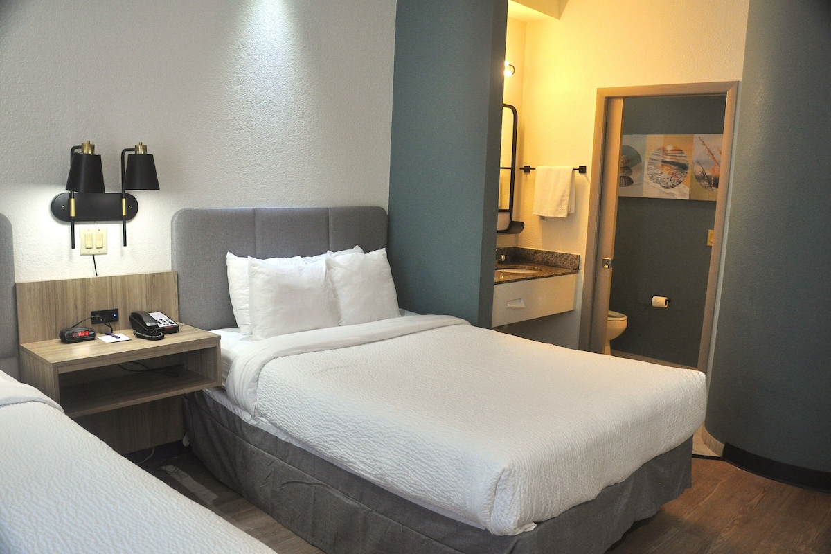 A hotel room with a bed, bedside table, lamp, and a bathroom visible through an open door.
