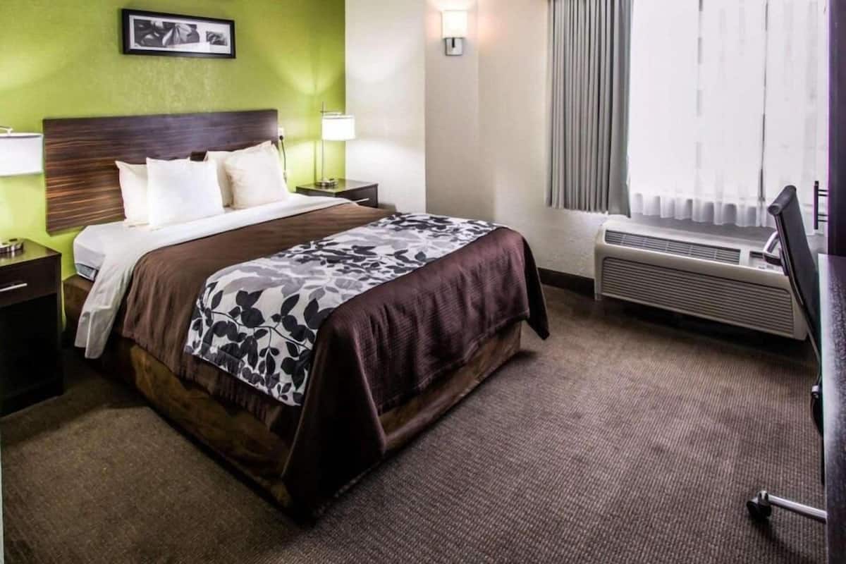 A hotel room with a bed, nightstands, a desk, a lamp, a window with curtains, and a wall-mounted air conditioner.