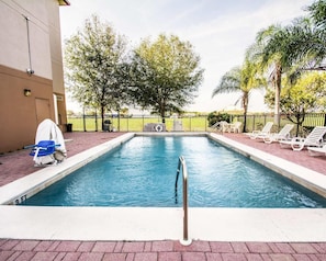 A rectangular swimming pool with a metal ladder, surrounded by a brick walkway, trees, and white lounge chairs.