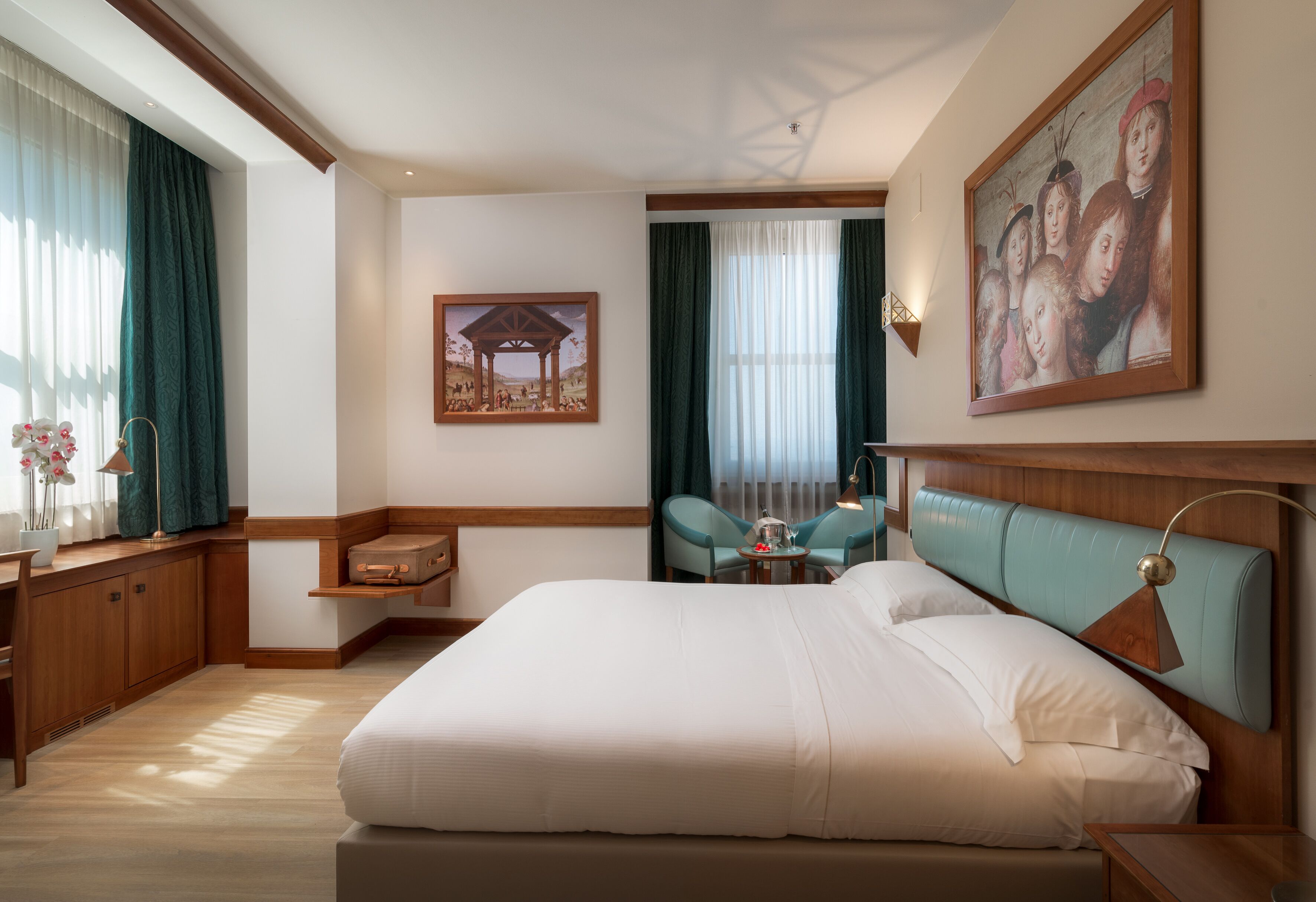 Habitación de hotel con una cama grande, un escritorio con silla, una mesita con dos sillas, un cuadro en la pared y una ventana con cortinas.