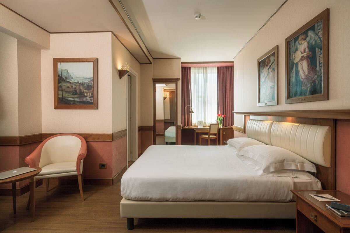 A hotel room with a large bed, a chair, a desk, and two paintings on the wall.