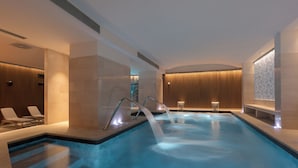 A modern indoor swimming pool with clear water, a sunken seating area, and a view of the cityscape through the windows. - Hotel Sangallo Palace (Perugia)