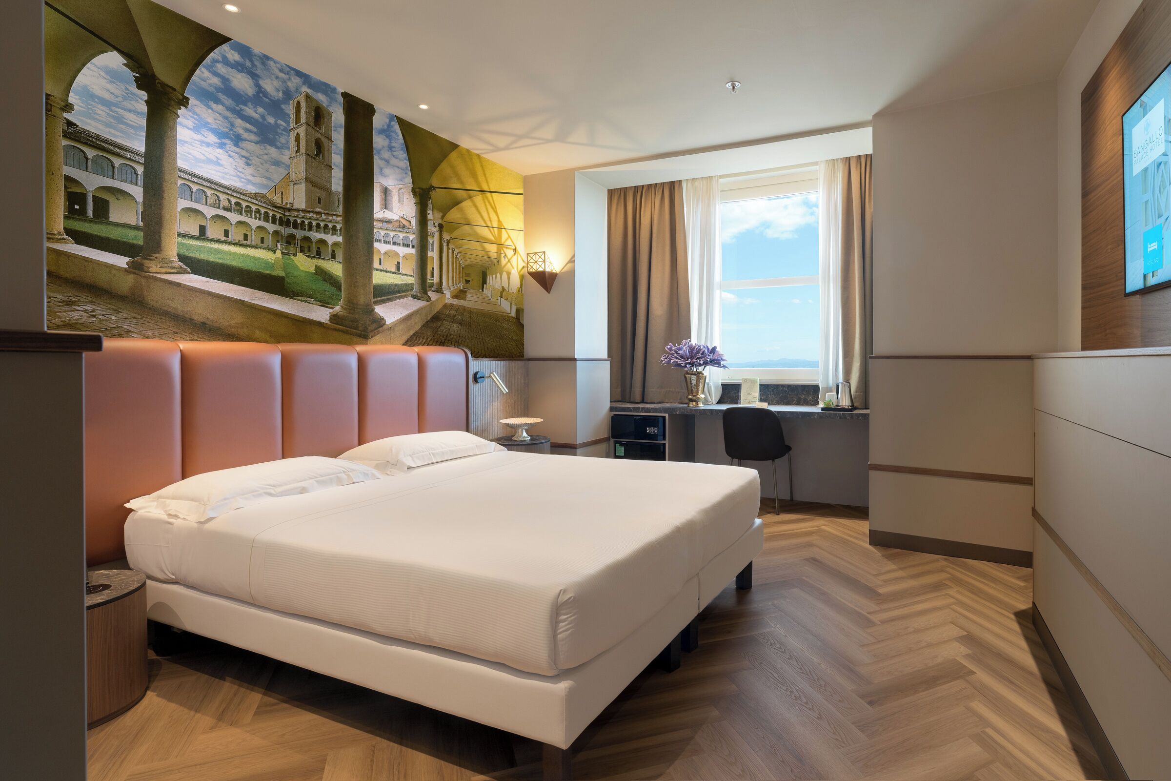Ein Hotelzimmer mit Bett, Schreibtisch und einem Wandbild eines Gebäudes.