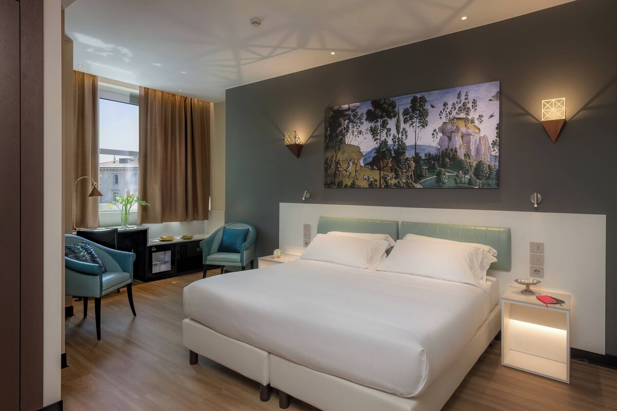 A modern hotel room with a large bed, a desk with a chair, a painting on the wall, and a nightstand with a lamp.