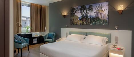 A modern hotel room with a large bed, a desk with a chair, a painting on the wall, and a nightstand with a lamp.