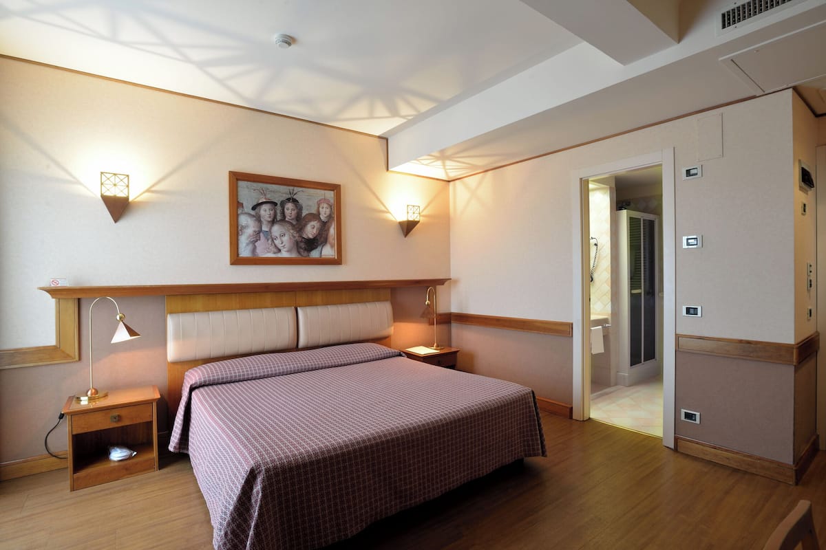 Un dormitorio con cama, mesitas de noche, un cuadro, apliques de pared y una puerta que da a un baño.