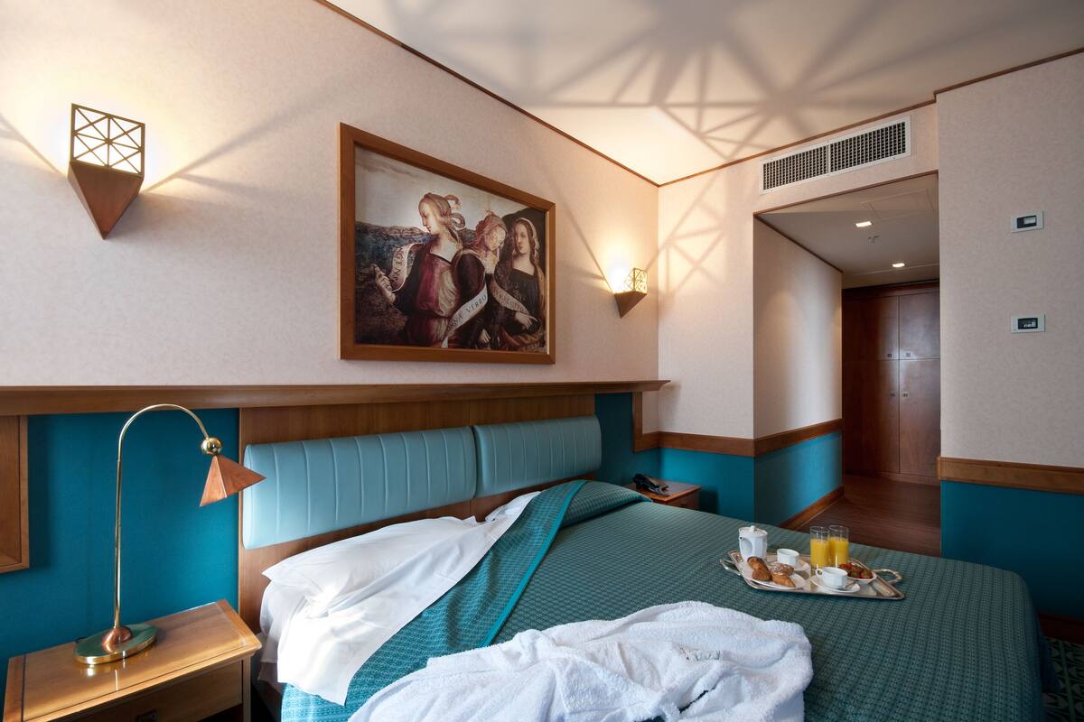 A hotel room with a bed, a bedside table, a lamp, a painting on the wall, and a tray with breakfast items.