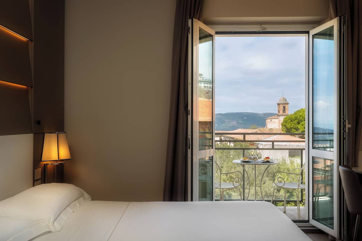 A hotel room with a bed, a nightstand with a lamp, and a balcony with a table and chairs.