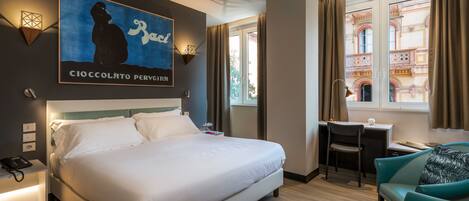 A hotel room with a large bed, a desk, and a chair. There is a painting on the wall and a window with curtains.