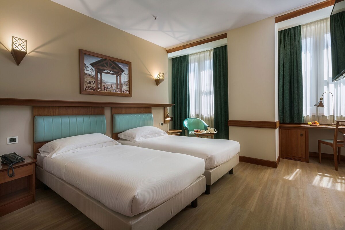 A hotel room with two beds, a desk, a chair, a telephone, and a painting on the wall.