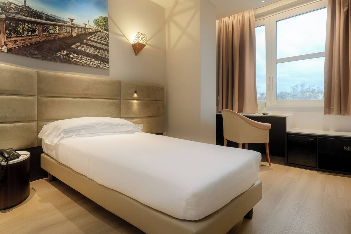 A hotel room with a bed, a desk, a chair, a nightstand, a wall-mounted lamp, and a window with curtains.