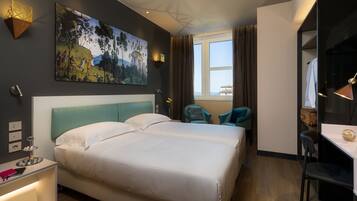 A modern hotel room with a large bed, a desk, and a chair. There is a painting on the wall and a window with curtains.