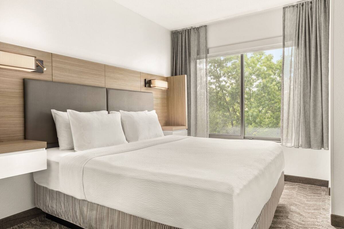 A modern hotel room with a large bed, a wooden headboard, a bedside table, and a window with curtains.