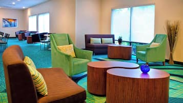 A hotel lobby with green armchairs, a brown sofa, and wooden coffee tables. There are multiple windows providing natural light.