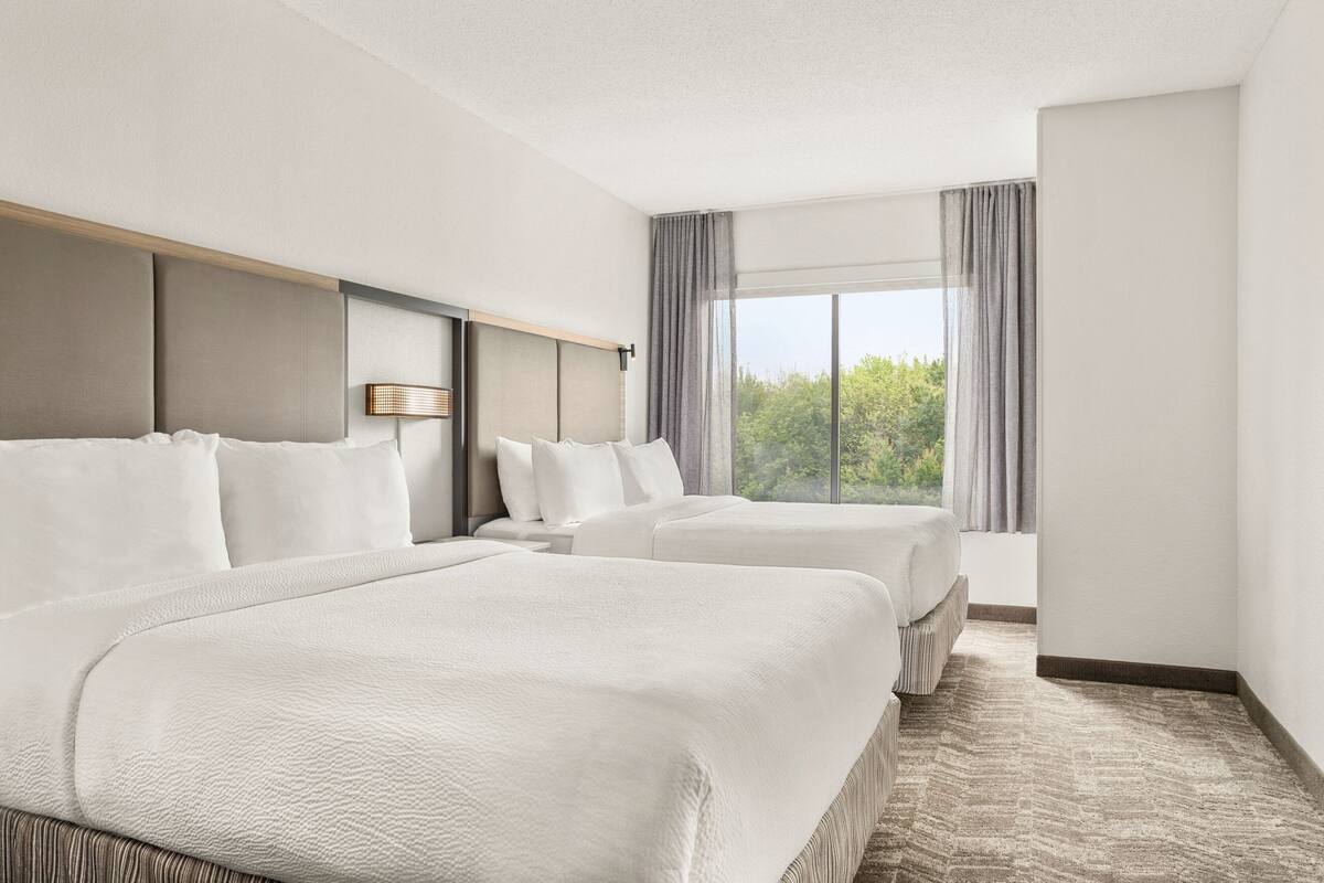 A hotel room with two beds, a large window with curtains, and a view of greenery outside.