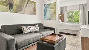 Une chambre d’hôtel moderne avec un canapé gris, une table basse en bois, un lit avec une literie blanche et de grandes fenêtres donnant sur un paysage verdoyant.