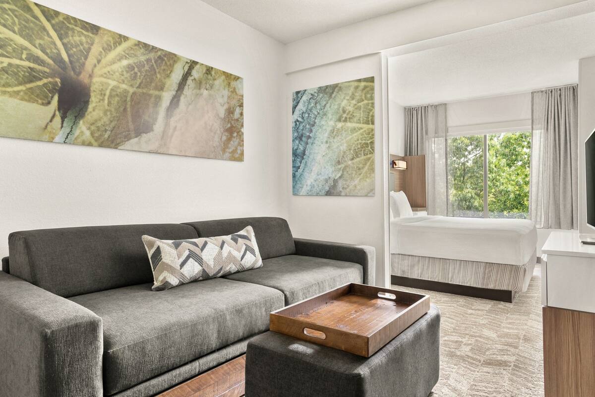 A modern hotel room with a gray sofa, a wooden coffee table, a bed with white bedding, and large windows offering a view of greenery.