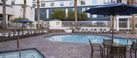 A pool area with lounge chairs and umbrellas, surrounded by palm trees and a hotel building in the background.
