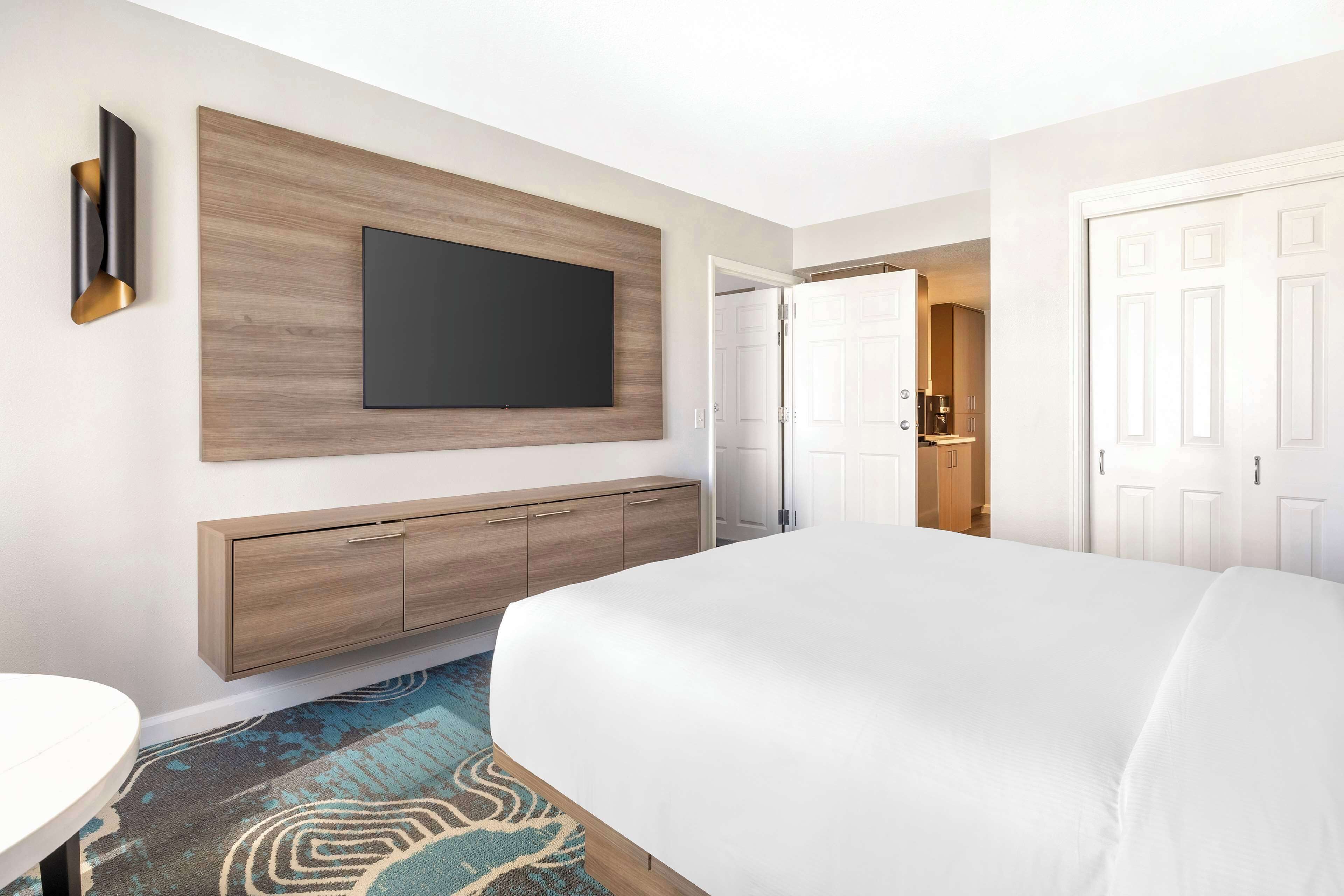 Una camera d'albergo con un letto grande, una TV a schermo piatto montata su un pannello di legno, un mobiletto in legno con cassetti e un tappeto decorato.