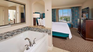 A hotel room with a large bathtub, a bed with a teal bedspread, a television on a wooden dresser, and a view of the city through the window.