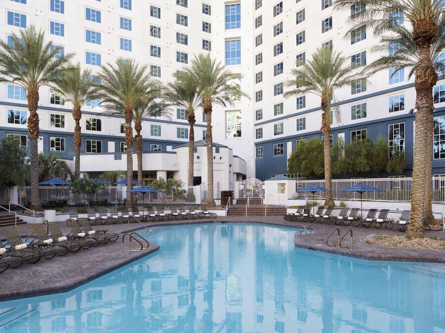 A hotel pool area with multiple lounge chairs and palm trees.