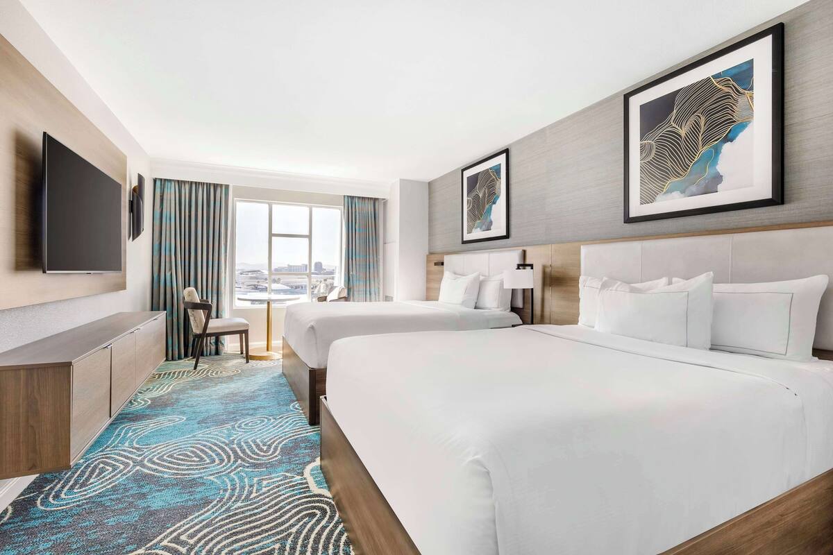 Habitación de hotel con dos camas, televisión, escritorio y vista a la ciudad.