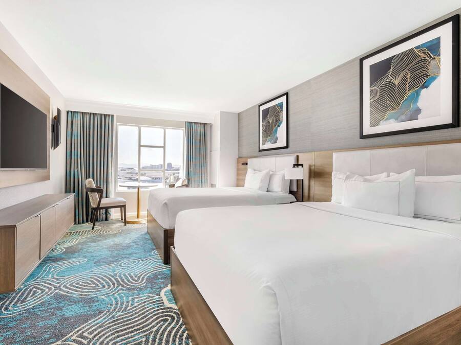 Habitación de hotel con dos camas, televisión, escritorio y vista a la ciudad.