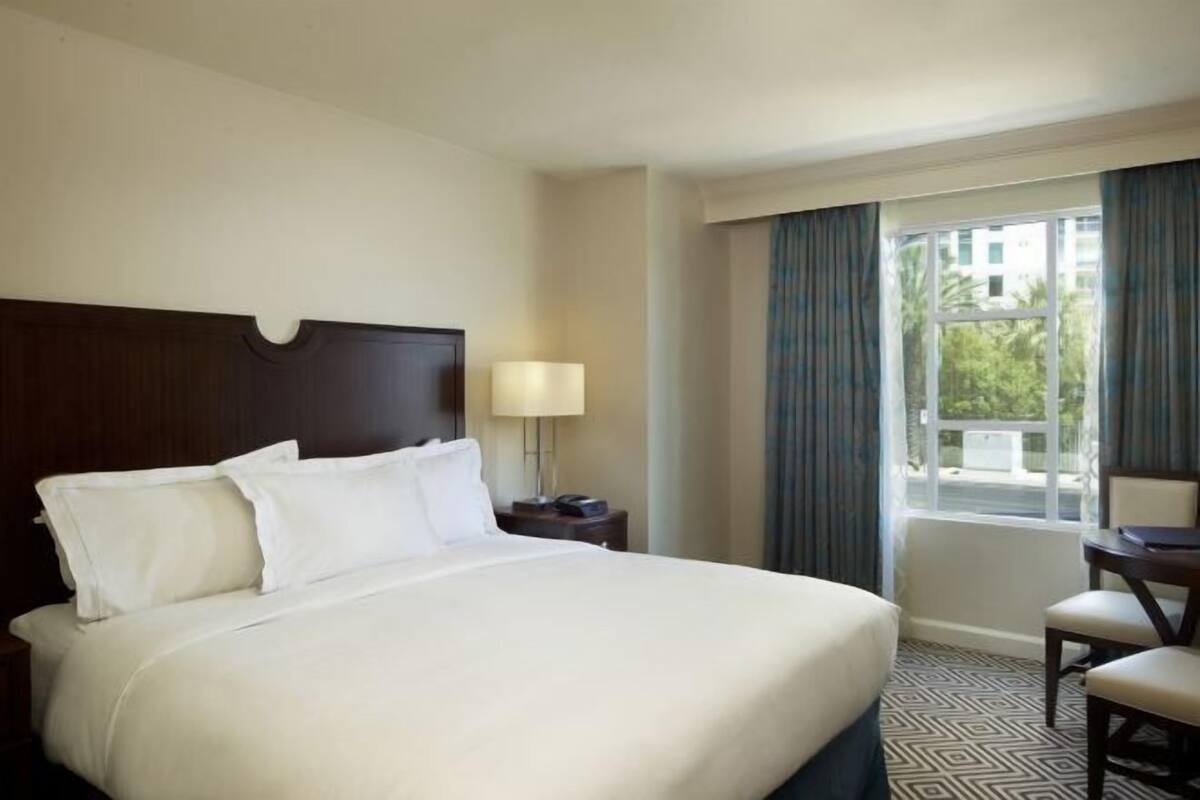 Habitación de hotel con una cama grande, una mesita de noche con lámpara, una silla y una ventana con cortinas.