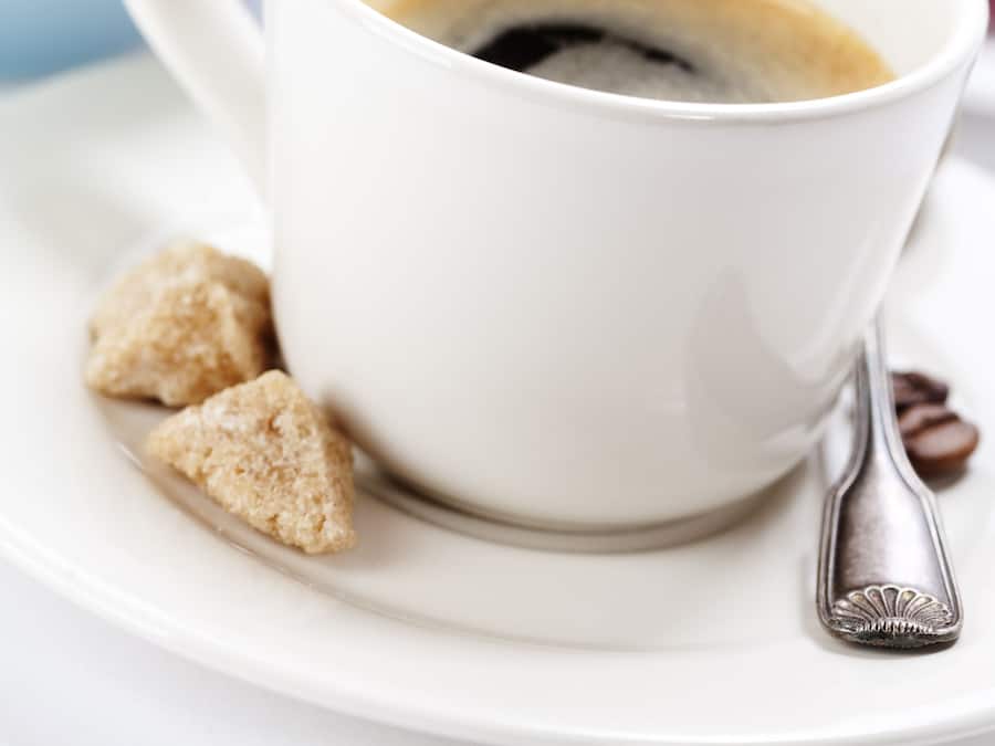 Una tazza di caffè bianco su un piattino, con un cucchiaino, accompagnata da una ciotola di frutta e un croissant.